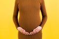 Close up of pregnant woman touching her abdomen at yellow background. Future mother is wearing brown dress. Expecting of a baby.