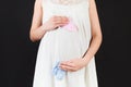 Close up of pregnant woman in white dress holding baby pink and blue socks against her belly at black background. Is it a boy or a Royalty Free Stock Photo