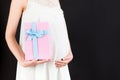 Close up of pregnant woman in white dress holding pink spotted gift box at black background. Expecting a baby girl. Copy space Royalty Free Stock Photo