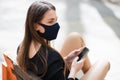 Close-up of a pretty brunette`s face, in a black protective mask. Woman sitting on a bench in the park with a cellphone in her Royalty Free Stock Photo