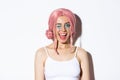 Close-up of pretty coquettish girl in pink wig, winking at camera and smiling, standing over white background