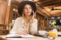 Close up of a pretty girl in hat sitting at the cafe table indoors, talking on mobile phone, taking notes in a notebook, Royalty Free Stock Photo