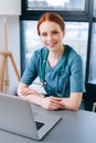 Close-up of pretty smiling young female doctor in blue green medical uniform sitting at desk with laptop on background Royalty Free Stock Photo