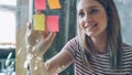 Close-up of pretty young woman with blond hair sticking colored memos on glass board in modern office. She is looking at