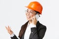 Close-up of professional smiling asian female entrepreneur at factory, chief engineer in safety helmet and suit, talking Royalty Free Stock Photo