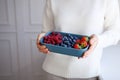 Close-up of raspberries, strawberries, blueberries in blue bowl in hands of woman in white sweater at light background. Copy space