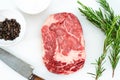 Close-up of raw Rib eye steak, peppercorns, salt and a few sprigs of fresh rosemary on a white surface. Food concept. Aerial view