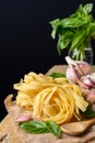 Close-up of raw tagliatelle nests on burlap with wooden spoon, basil and garlic, selective focus, on rustic table and black backgr