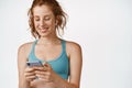 Close up of redhead fitness girl listening music in headphones and looking at smartphone, using sport app, standing over Royalty Free Stock Photo