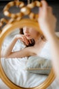 Reflection of young lady in wedding dress adorned with feathers holding gold-rimmed mirror, resting on blue pillow. Royalty Free Stock Photo