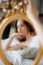 Reflection of brunette rest on bed in wedding gown with feathers leaning head on hand and holding gold-rimmed mirror. Royalty Free Stock Photo