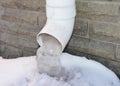 A close-up of a roof gutter downpipe, downspout with frozen water, icicles near the house foundation in winter