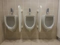 Close up of a row of three white ceramic urinals fixed on a beige tile wall separated by privacy walls