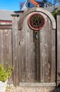 Close-up of rustic wooden gate with decorative round wrought iron window and a planting trowel for a knocker with two story house