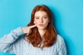 Close-up of sassy teen girl with red hair, pointing at her cheek and staring at camera, standing against blue background