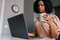 Close-up selective focus shot of African American business woman typing on laptop looking to device screen and drinking Royalty Free Stock Photo