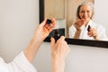 Close up of a senior woman`s hands holding a dropper and a bottle with a liquid facial treatment in bathroom