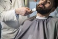 Close up of a serious brown haired businessman having his beard combed and trimmed in a barber shop.