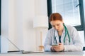 Close-up of serious female doctor wearing white coat using mobile phone while sitting at desk with laptop on background Royalty Free Stock Photo