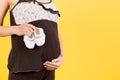 Close up of shoes for baby girl in pregnant woman`s hands at yellow background. Future mother is wearing brown pajamas. Motherhoo