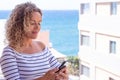 Close up shot of beautiful curly woman smiling while using phone. Blue sea in background. Female model with copy space Royalty Free Stock Photo