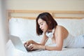 Close up shot of beautiful and sexy young adult asian woman in white dress lying on bed in bedroom while using computer and Royalty Free Stock Photo