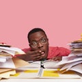 Close up shot black dark skinned male wonk covers mouth with hand, looks through piles of books and papers, wears