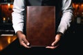 A black waiter\'s hands holding a blank brown leather-bound menu facing towards the camera - Template mock-up