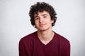 Close up shot of curly haired European teenager with little beard, wears maroon shirt. Young smiling male stands with lowered Royalty Free Stock Photo