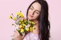 Close up shot of dark haired woman with healthy skin, closes eyes with enjoyment, holds pretty flowers closely, poses over rosy