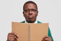 Close up shot of displeased young African American man with gloomy facial expression, purses lips, holds spiral notepad