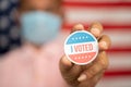 Close up shot man in medical mask showing I voted Sticker and putting on shirt with US flag as background - concept of US election Royalty Free Stock Photo