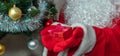 Close up shot of Santa Claus holding a red gift box in both hands. Christmas tree with red, white, and golden balls, white tinsel Royalty Free Stock Photo