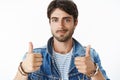 Close-up shot of satisfied and happy young attractive male with blue eyes and bristle in denim jacket raising thumbs up Royalty Free Stock Photo