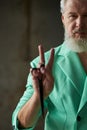 Close up shot of stylish middle aged man with beard wearing colorful outfit looking at camera, showing peace sign while Royalty Free Stock Photo