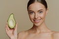 Topless young Caucasian female model has dark combed hair smiles gently holds half of avocado stands shirtless indoor Royalty Free Stock Photo