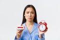 Close-up shot of upset silly asian girl on diet, showing clock and tasty piece of cake, pouting as want to eat dessert