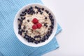 Close up shot of white bowl with porridge being on blue kitchen towel, oatmeal decorated with raspberry and blueberry at top,