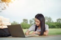 Young female university student writes a note and assignment with a laptop beside outside the campus with blur green tree backgrou