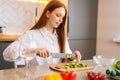 Close-up side view of attractive young redhead woman cutting fresh cucumber cooking food salad sitting at table in Royalty Free Stock Photo