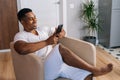 Close-up side view of cheerful African-American man sitting at armchair and using mobile phone. Royalty Free Stock Photo