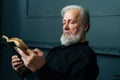 Close-up side view of senior gray-haired aged male reading paper book sitting on chair in dark living room, relax and Royalty Free Stock Photo