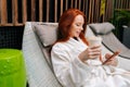 Close-up side view of smiling young woman in white bathrobe relaxing with mobile sitting on deck chair by poolside and Royalty Free Stock Photo