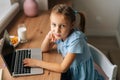 Close-up side view of tired elementary child girl using laptop sitting at home table with snack by window, looking at Royalty Free Stock Photo