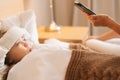 Close-up side view of worried mother sitting on bed beside sick daughter with high fever and calling on phone to doctor