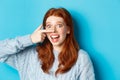 Close-up of silly redhead girl showing piggy nose, making funny grimaces and smiling, standing against blue background