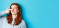 Close-up of silly redhead girl showing piggy nose, making funny grimaces and standing against blue background