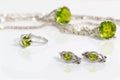 Close-up silver earrings and small ring with peridot on background pendant, chain and big ring on white acrylic desk.