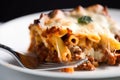 Close-up of a single baked ziti, cut in half, with a fork on a white plate, showcasing the layers of pasta, cheese, and meat