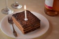 Slice of a layered chocolate cake with one candle and two glasses and bottle of wine